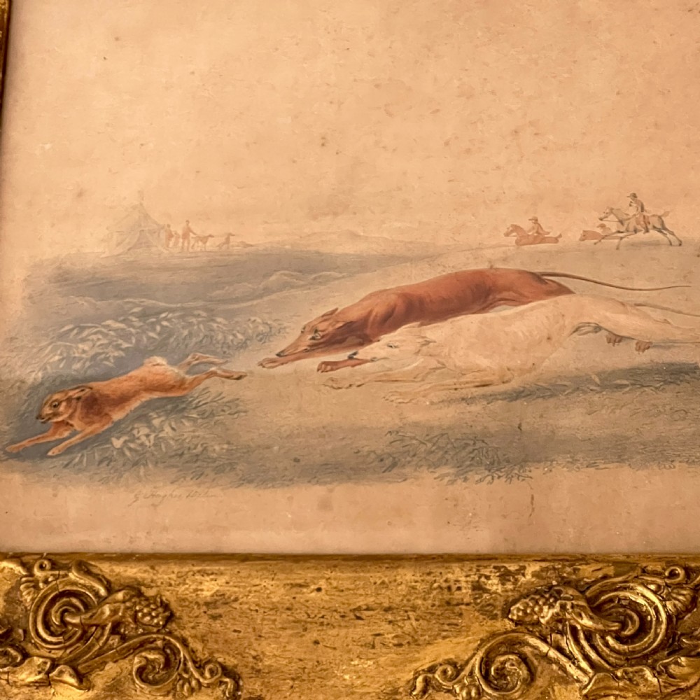 A 19th-century watercolour painting depicting two greyhounds pursuing a hare across a field, housed in a wide, ornate gilt frame with acanthus corner details. View IV