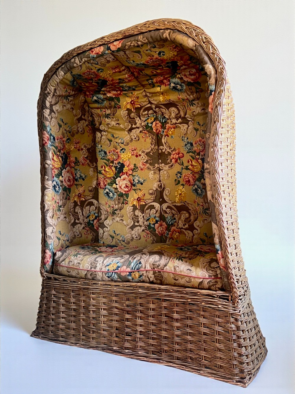 A high-backed woven wicker porters chair featuring a hooded canopy and floral upholstered linen interior. View V