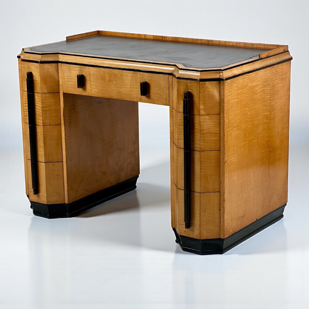 An Art Deco satin birch writing desk featuring a black leather top, stepped geometric corners, dark trim, and twin pedestals with drawers.