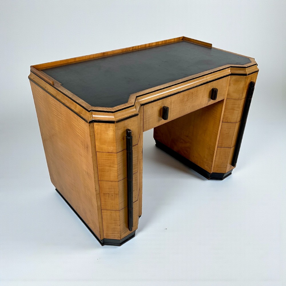 An Art Deco satin birch writing desk featuring a black leather top, stepped geometric corners, dark trim, and twin pedestals with drawers. View II