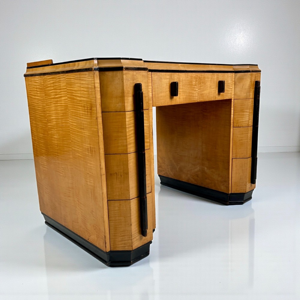 An Art Deco satin birch writing desk featuring a black leather top, stepped geometric corners, dark trim, and twin pedestals with drawers. View IV