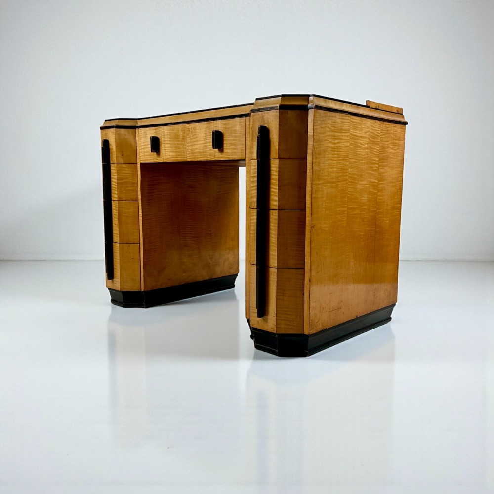 An Art Deco satin birch writing desk featuring a black leather top, stepped geometric corners, dark trim, and twin pedestals with drawers. View V