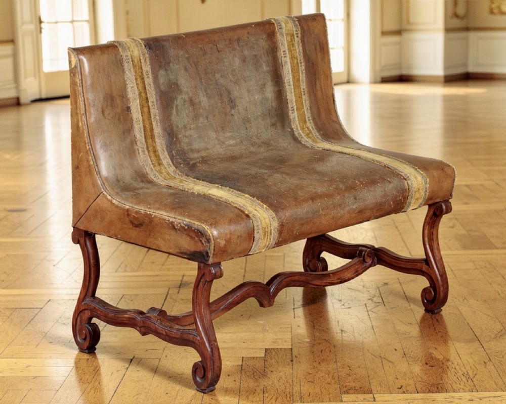 An antique French leather banquette featuring patinated brown leather upholstery with two vertical velvet decorative bands, supported by a carved wooden base with scrolled legs.