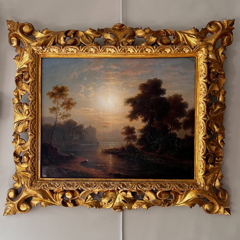 An antique oil painting on canvas depicting a tranquil riverscape framed by dense trees and a soft sky, enclosed within an ornate carved giltwood frame. View IV