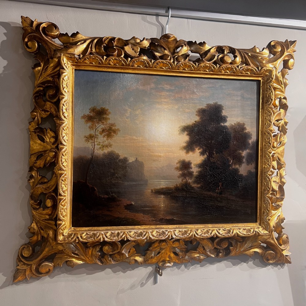 An antique oil painting on canvas depicting a tranquil riverscape framed by dense trees and a soft sky, enclosed within an ornate carved giltwood frame. View V