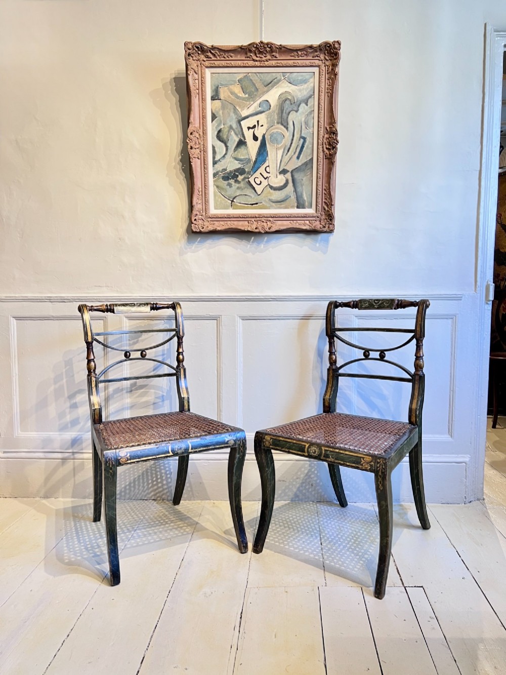 A pair of Regency period painted side chairs featuring woven cane seats, sabre legs, and gilt hand-painted detailing on a dark frame. View II