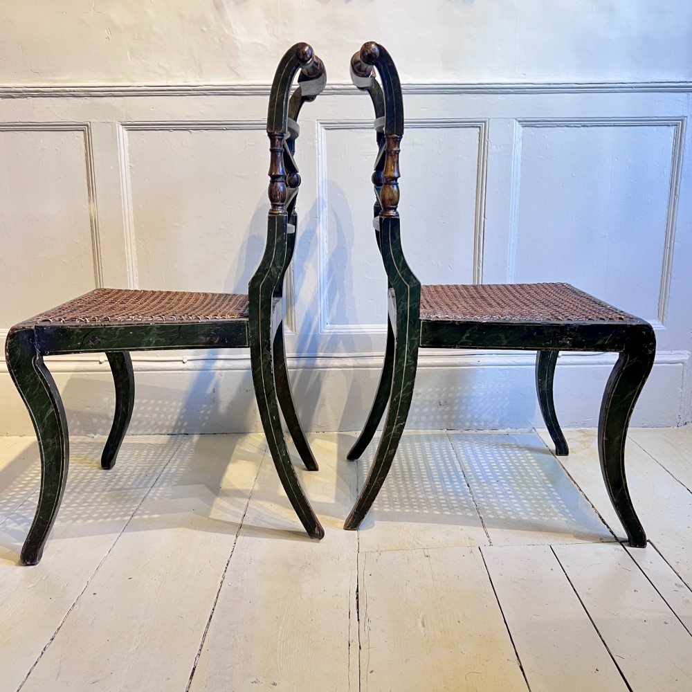 A pair of Regency period painted side chairs featuring woven cane seats, sabre legs, and gilt hand-painted detailing on a dark frame. View III