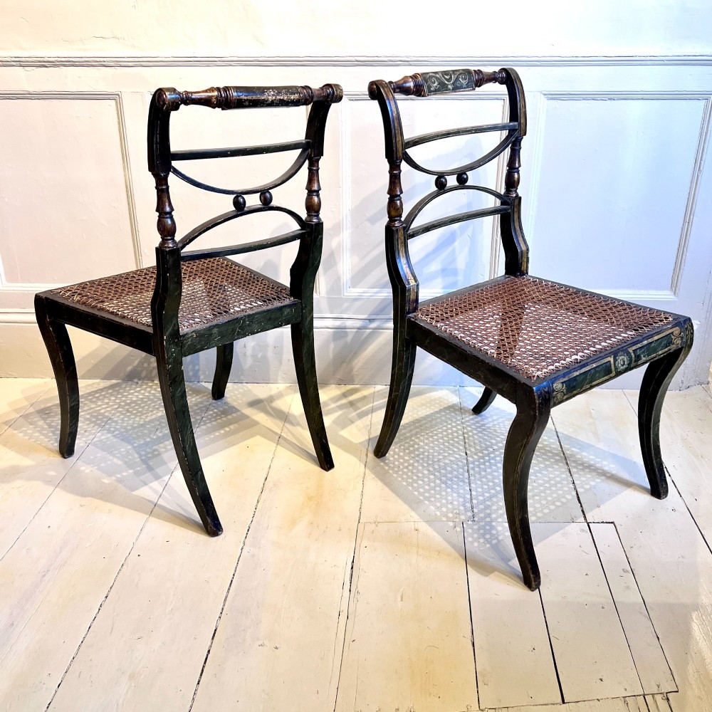 A pair of Regency period painted side chairs featuring woven cane seats, sabre legs, and gilt hand-painted detailing on a dark frame. View IV