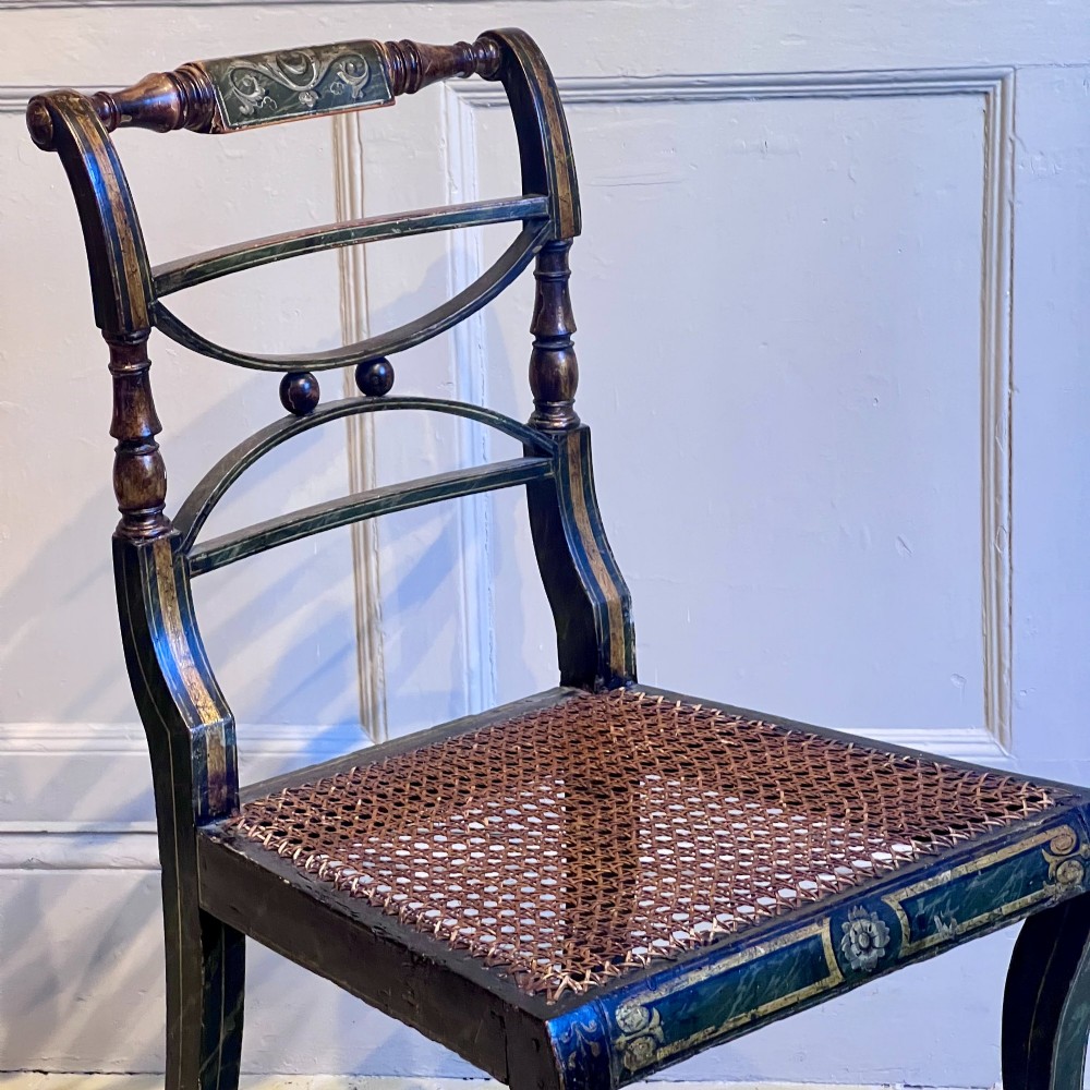 A pair of Regency period painted side chairs featuring woven cane seats, sabre legs, and gilt hand-painted detailing on a dark frame. View V