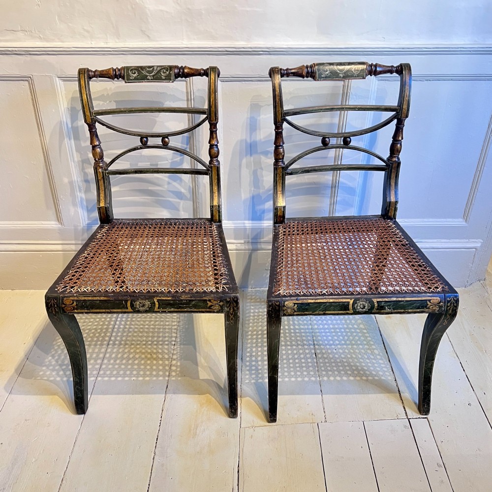 A pair of Regency period painted side chairs featuring woven cane seats, sabre legs, and gilt hand-painted detailing on a dark frame. View VII