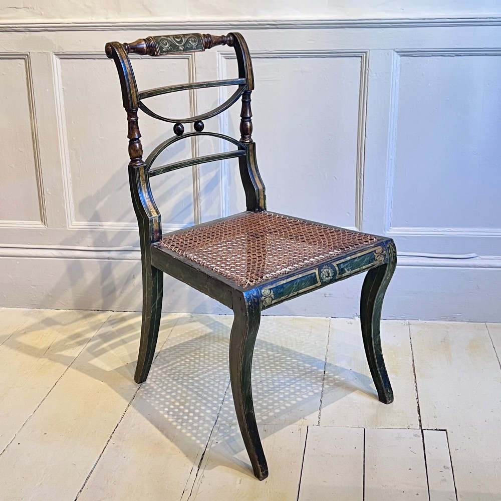 A pair of Regency period painted side chairs featuring woven cane seats, sabre legs, and gilt hand-painted detailing on a dark frame. View VIII