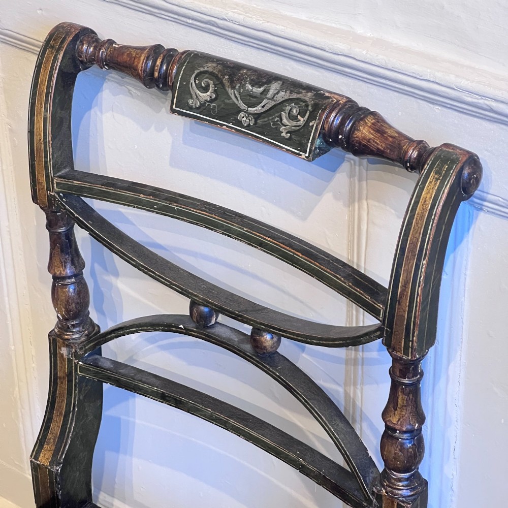 A pair of Regency period painted side chairs featuring woven cane seats, sabre legs, and gilt hand-painted detailing on a dark frame. View X