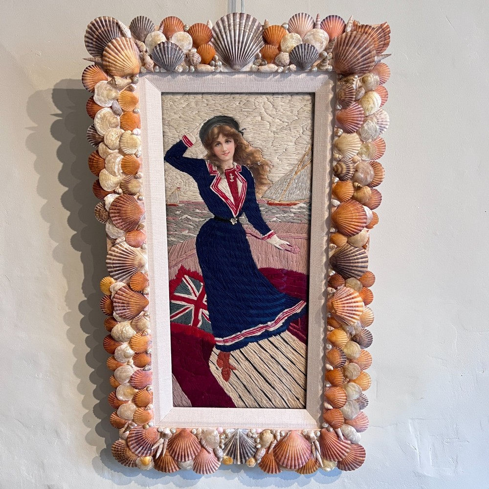 A framed Victorian woolwork picture depicting a sailor girl standing on a ship deck beside a Union Jack flag, enclosed within a rectangular frame decorated with natural sea shells. View II