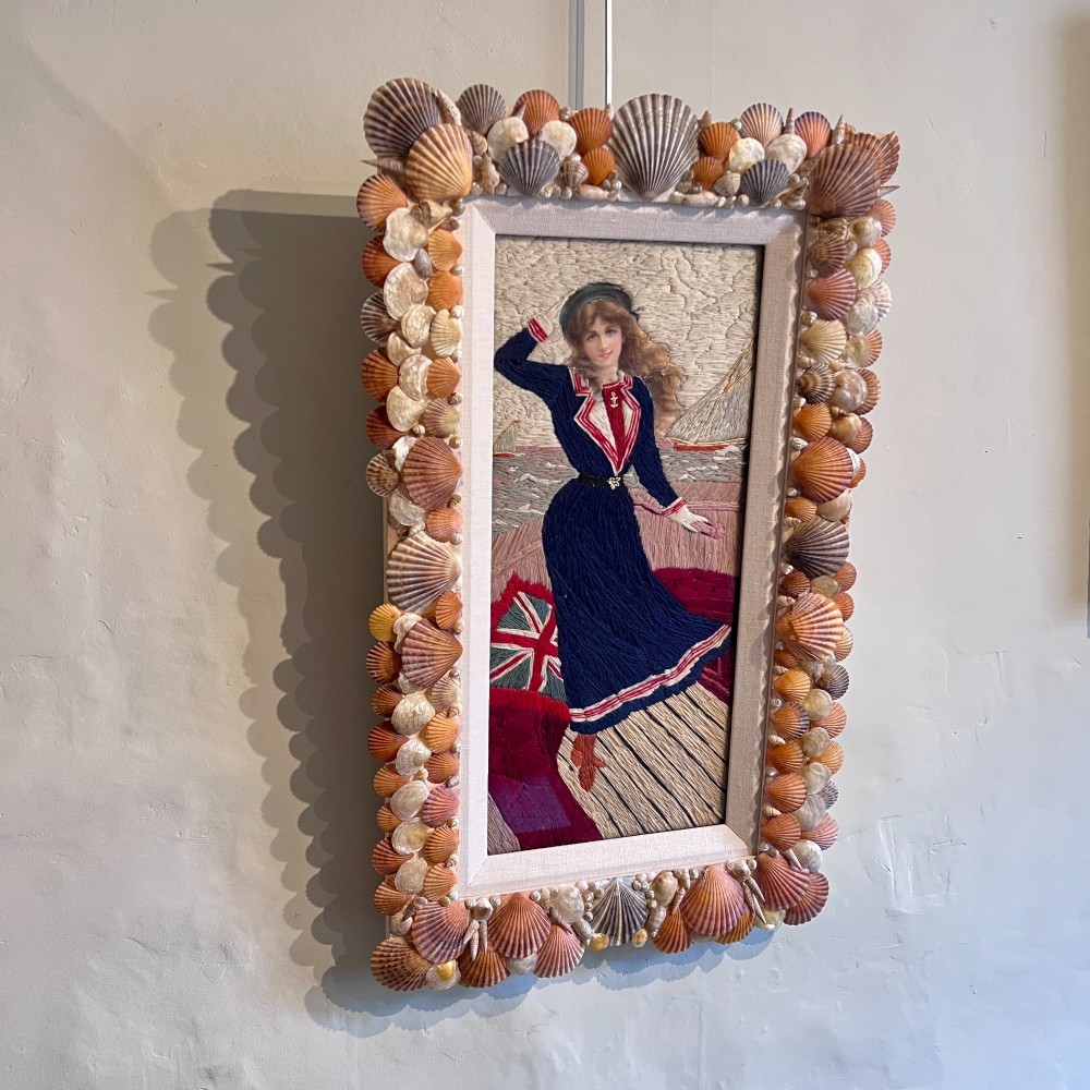 A framed Victorian woolwork picture depicting a sailor girl standing on a ship deck beside a Union Jack flag, enclosed within a rectangular frame decorated with natural sea shells. View III