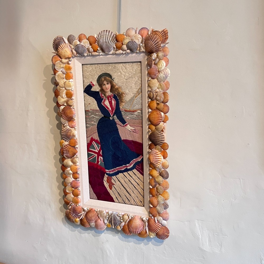 A framed Victorian woolwork picture depicting a sailor girl standing on a ship deck beside a Union Jack flag, enclosed within a rectangular frame decorated with natural sea shells. View IV