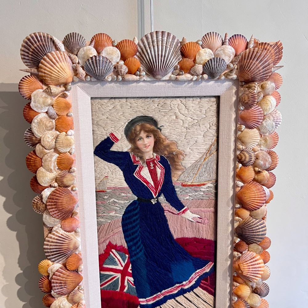 A framed Victorian woolwork picture depicting a sailor girl standing on a ship deck beside a Union Jack flag, enclosed within a rectangular frame decorated with natural sea shells. View V