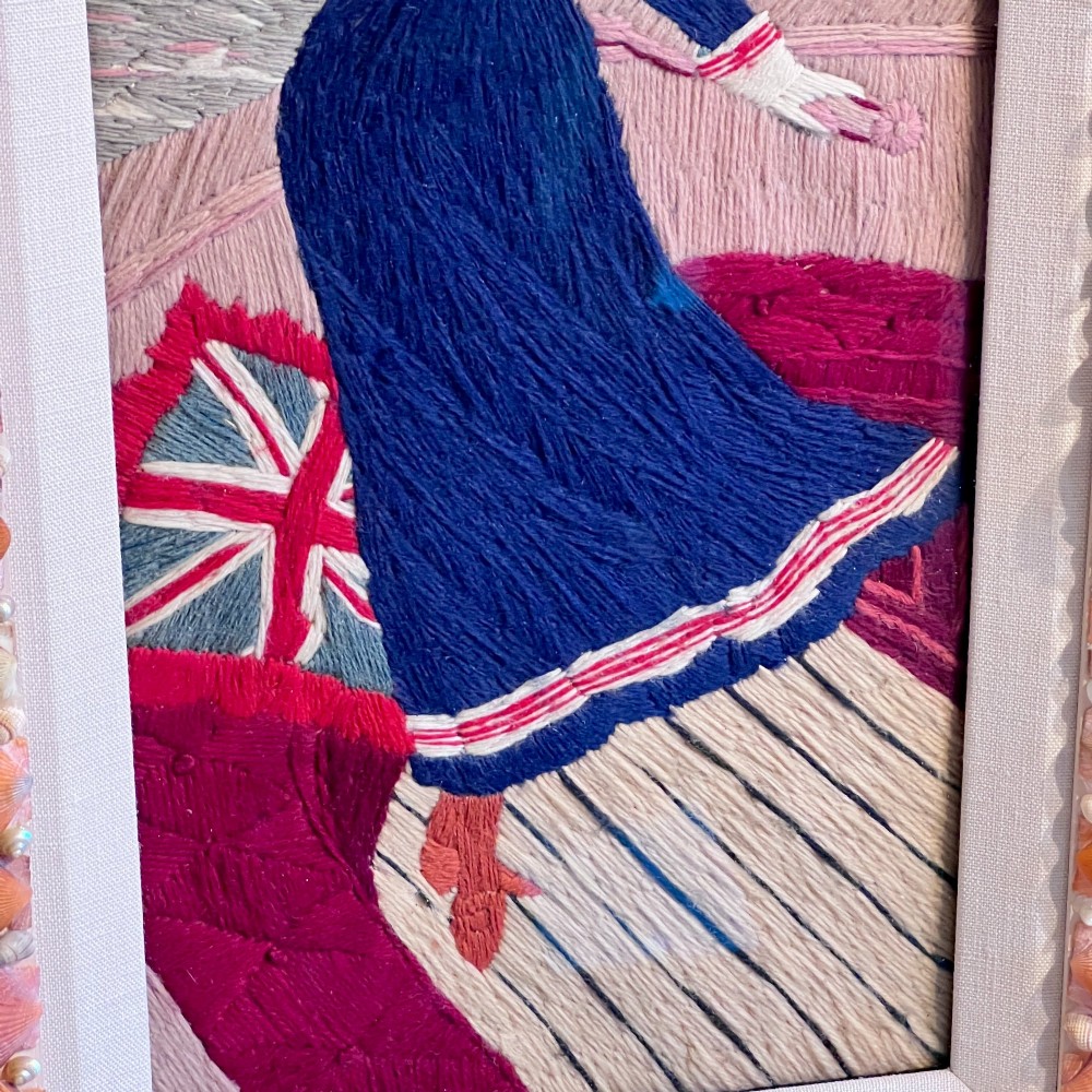 A framed Victorian woolwork picture depicting a sailor girl standing on a ship deck beside a Union Jack flag, enclosed within a rectangular frame decorated with natural sea shells. View VIII