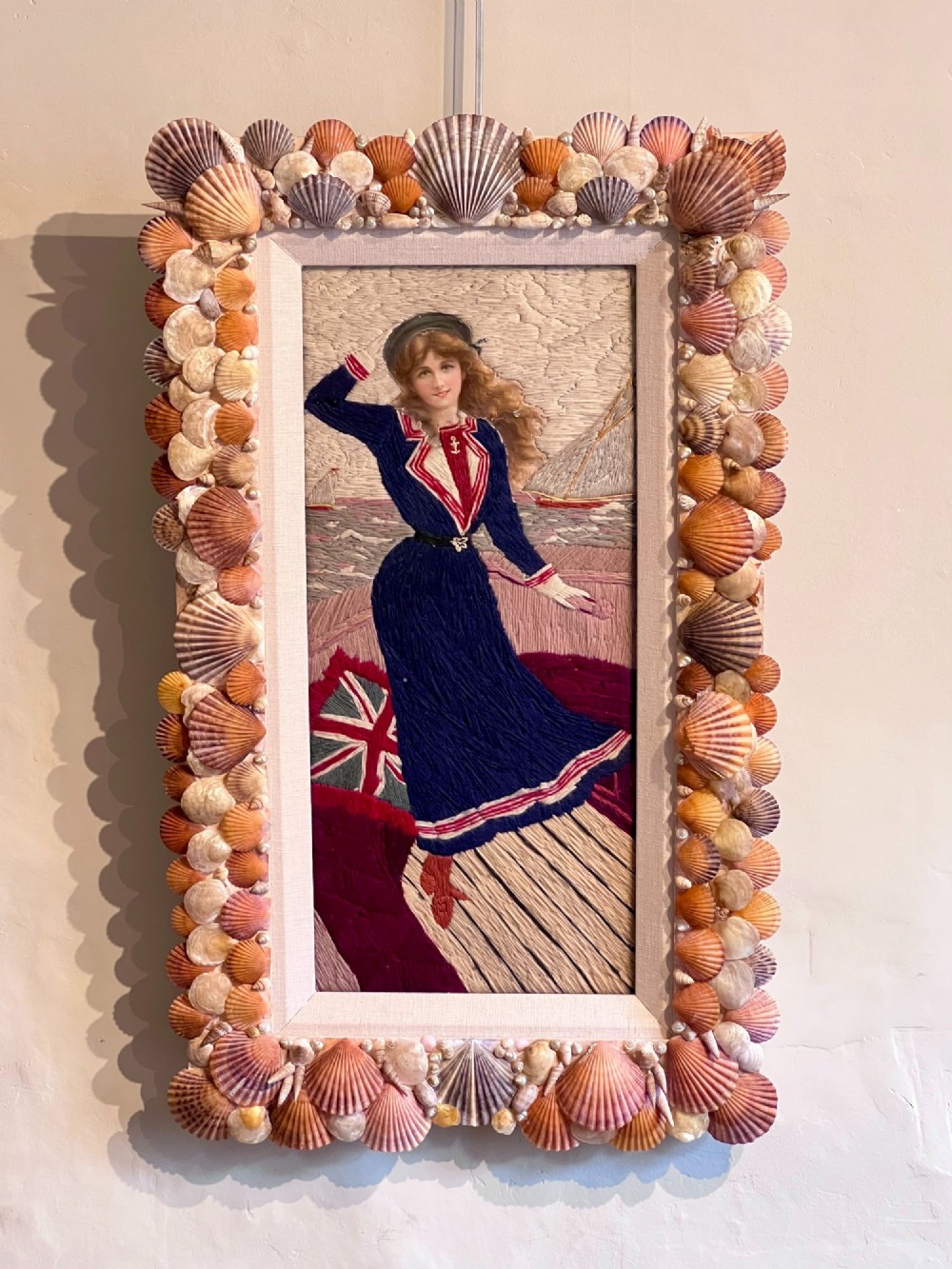 A framed Victorian woolwork picture depicting a sailor girl standing on a ship deck beside a Union Jack flag, enclosed within a rectangular frame decorated with natural sea shells. View IX