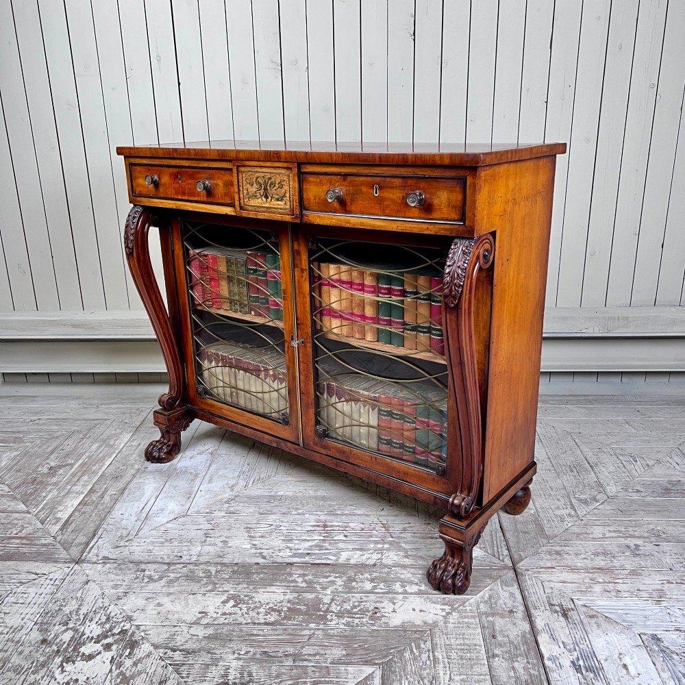A Regency mahogany low chiffonier bookcase featuring brass wire grille doors, carved scroll supports terminating in paw feet, and brass inlay.