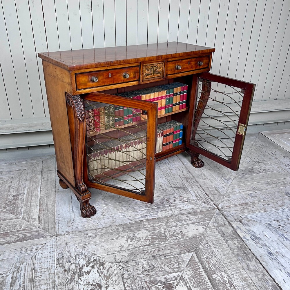 A Regency mahogany low chiffonier bookcase featuring brass wire grille doors, carved scroll supports terminating in paw feet, and brass inlay. View V