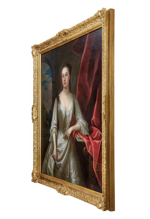 fig 2. A portrait depicting a woman in a silver satin dress seated next to draped red fabric, enclosed within an ornate carved gold frame.