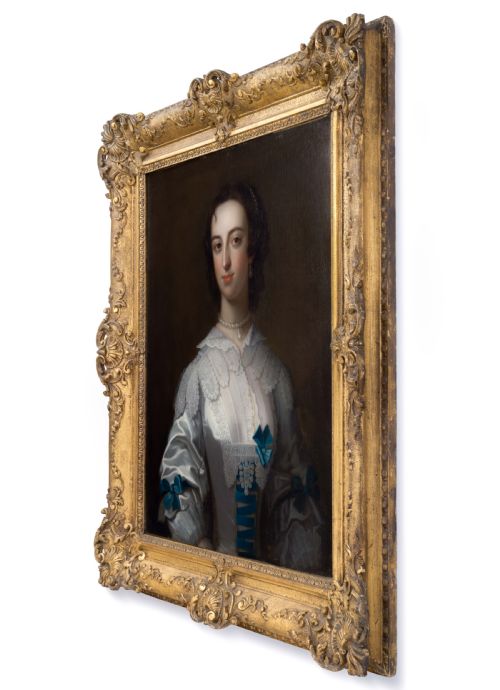 fig 1. A half-length mid-18th-century oil portrait of a lady wearing a white lace-trimmed silk gown decorated with blue ribbons, set within a carved giltwood frame.