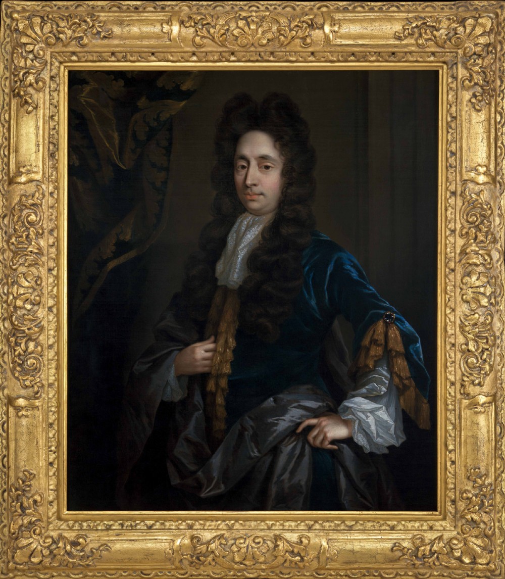 An oil painting of a 17th-century gentleman wearing a dark blue coat, full brown wig, and white lace cravat, set inside an ornate carved gilded frame.