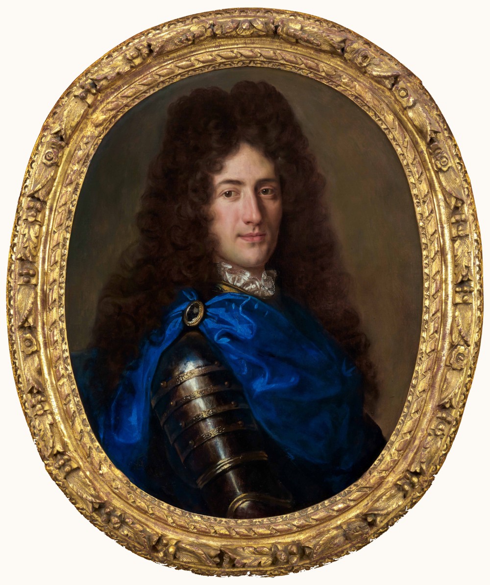 An oval oil painting depicting a gentleman wearing plate armour and an azure blue cloak with a jewelled brooch, set within a carved gold gilt frame.