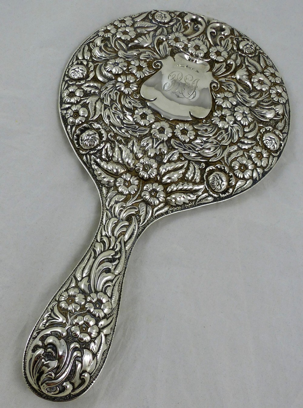 An Edwardian sterling silver hand mirror decorated with repousse floral scrollwork and a central engraved cartouche. View VI