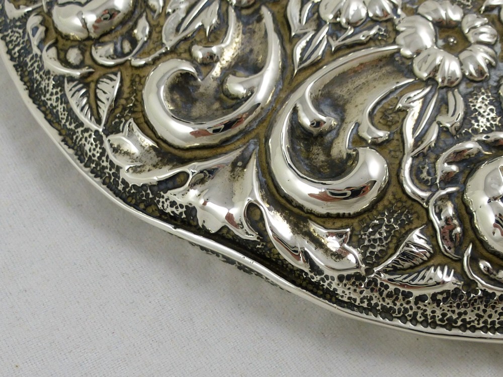 An Edwardian sterling silver hand mirror decorated with repousse floral scrollwork and a central engraved cartouche. View X