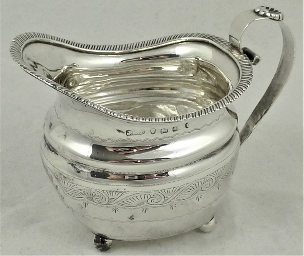 A Victorian sterling silver milk jug featuring gadrooned rim details, engraved geometric scrollwork, a shaped handle, and small ball feet.
