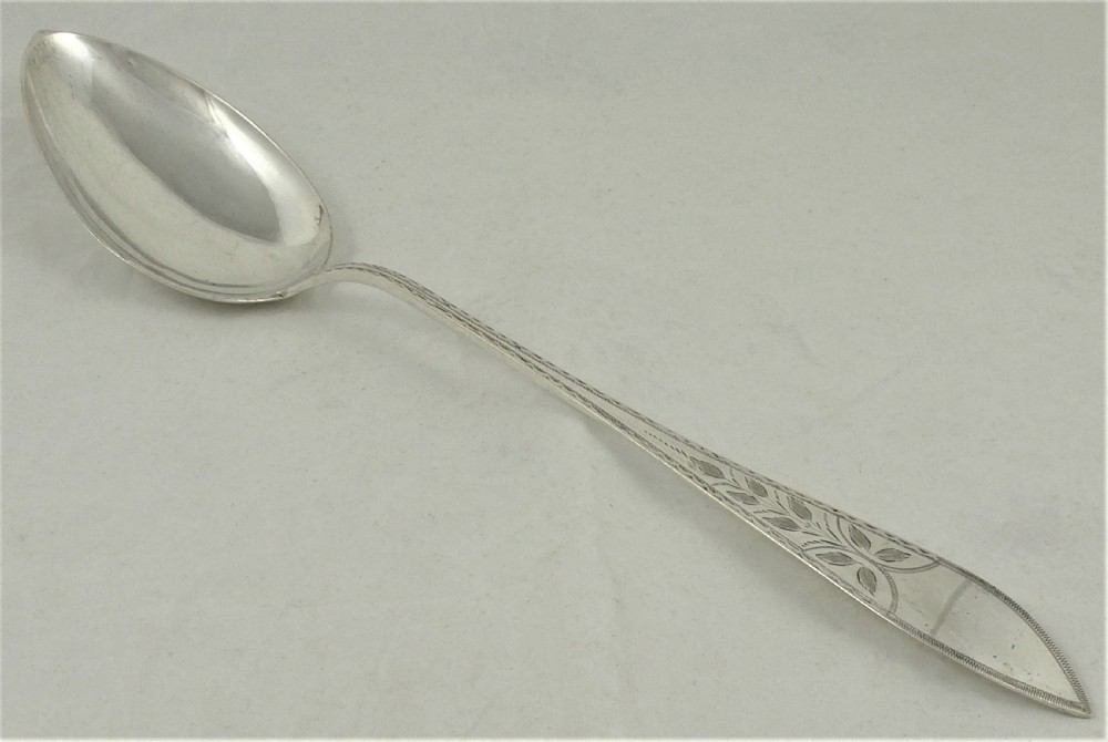An antique Danish silver serving spoon featuring a polished oval bowl and a tapered stem decorated with bright-cut foliage engraving.