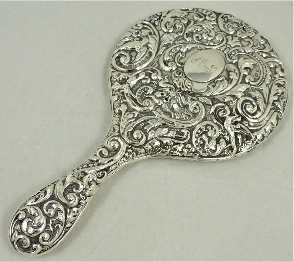 An ornate Edwardian sterling silver hand mirror featuring intricate scrolling foliage repousse decoration and a central engraved monogram.