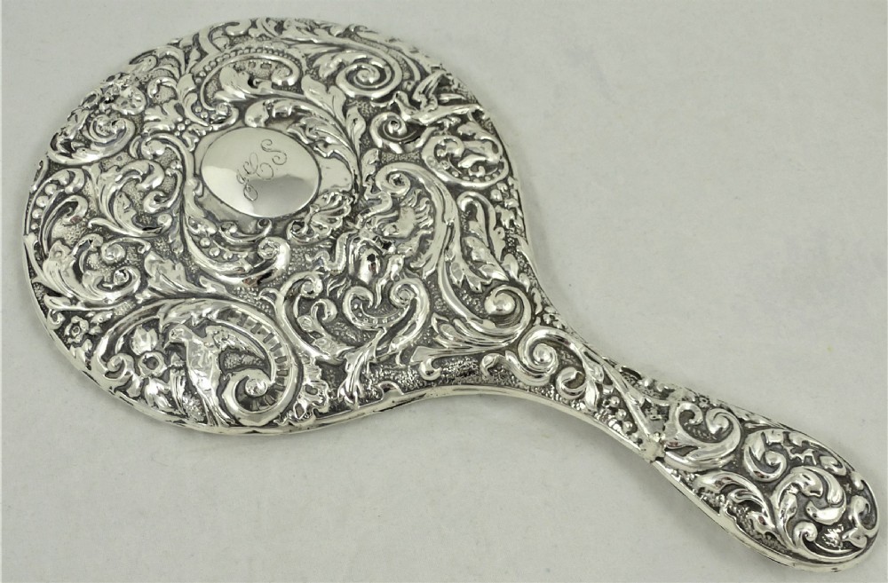 An ornate Edwardian sterling silver hand mirror featuring intricate scrolling foliage repousse decoration and a central engraved monogram. View V