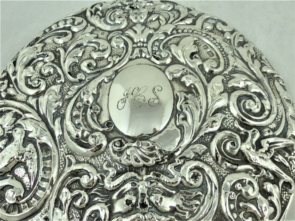An ornate Edwardian sterling silver hand mirror featuring intricate scrolling foliage repousse decoration and a central engraved monogram. View VI