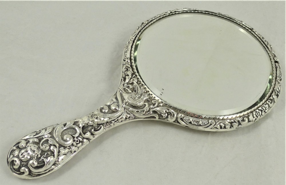 An ornate Edwardian sterling silver hand mirror featuring intricate scrolling foliage repousse decoration and a central engraved monogram. View X