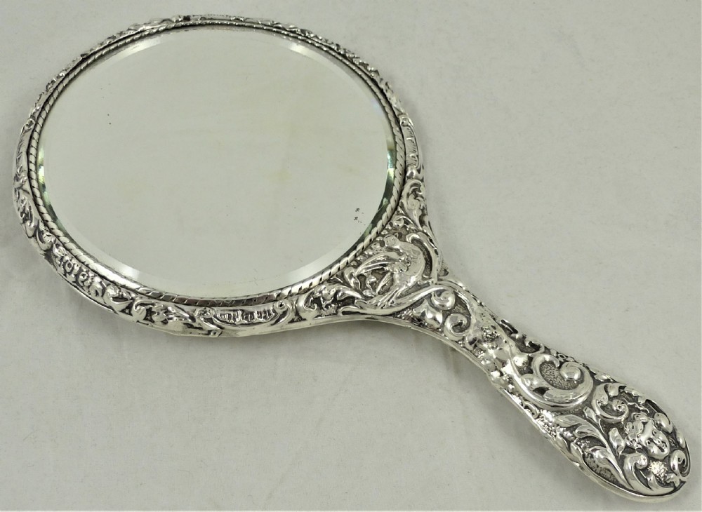An ornate Edwardian sterling silver hand mirror featuring intricate scrolling foliage repousse decoration and a central engraved monogram. View XI