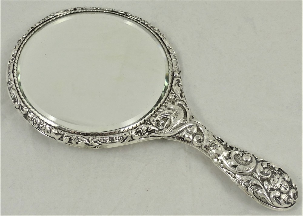 An ornate Edwardian sterling silver hand mirror featuring intricate scrolling foliage repousse decoration and a central engraved monogram. View XII