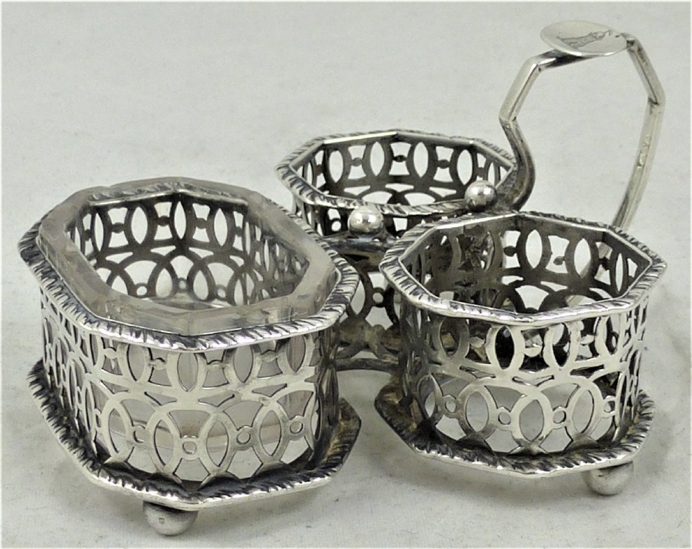 A Victorian silver cruet stand featuring three pierced octagonal compartments, small ball feet, an angled central loop handle, and one clear glass insert.