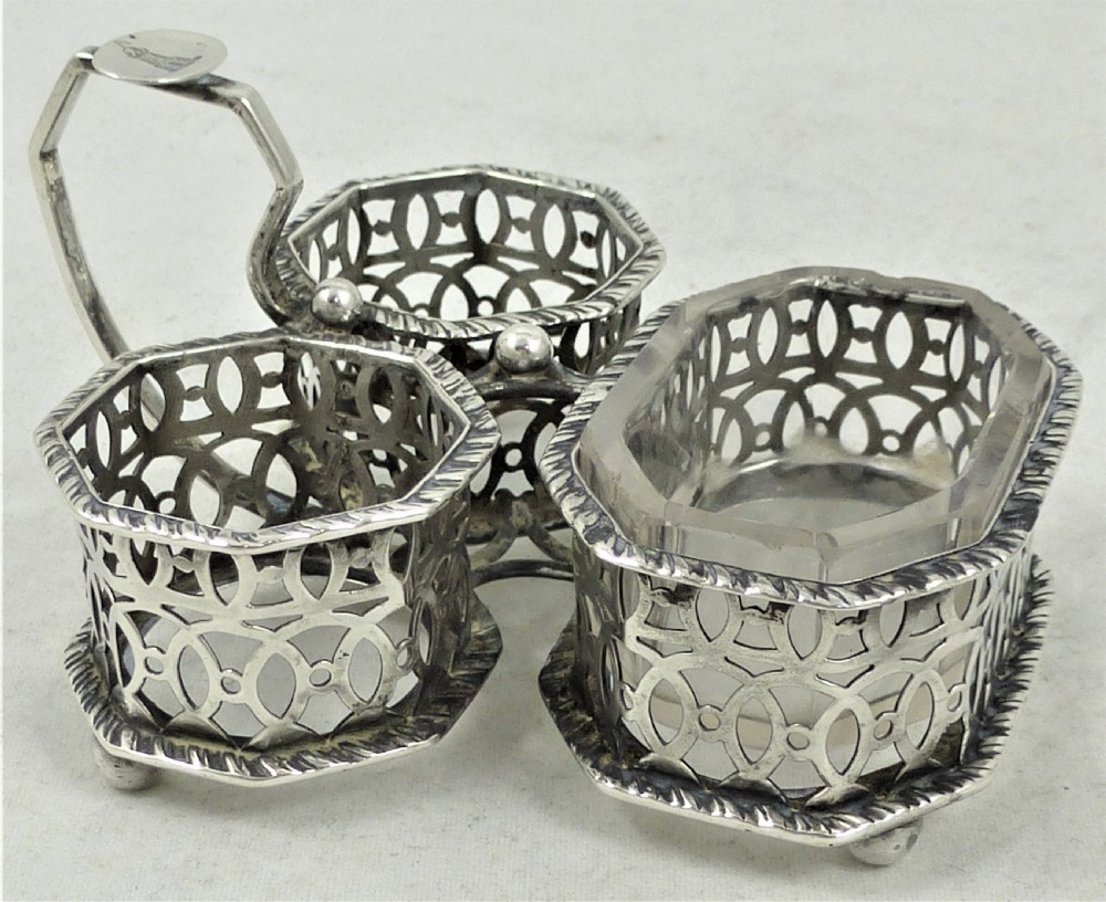A Victorian silver cruet stand featuring three pierced octagonal compartments, small ball feet, an angled central loop handle, and one clear glass insert. View V