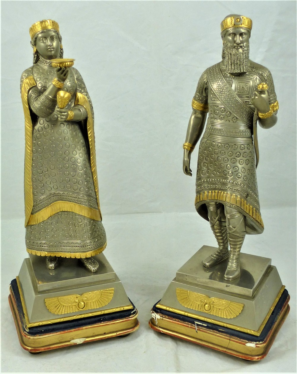 A pair of silvered and gilt metal statuettes depicting an ancient Assyrian king and queen standing on stepped plinths adorned with winged sun motifs.