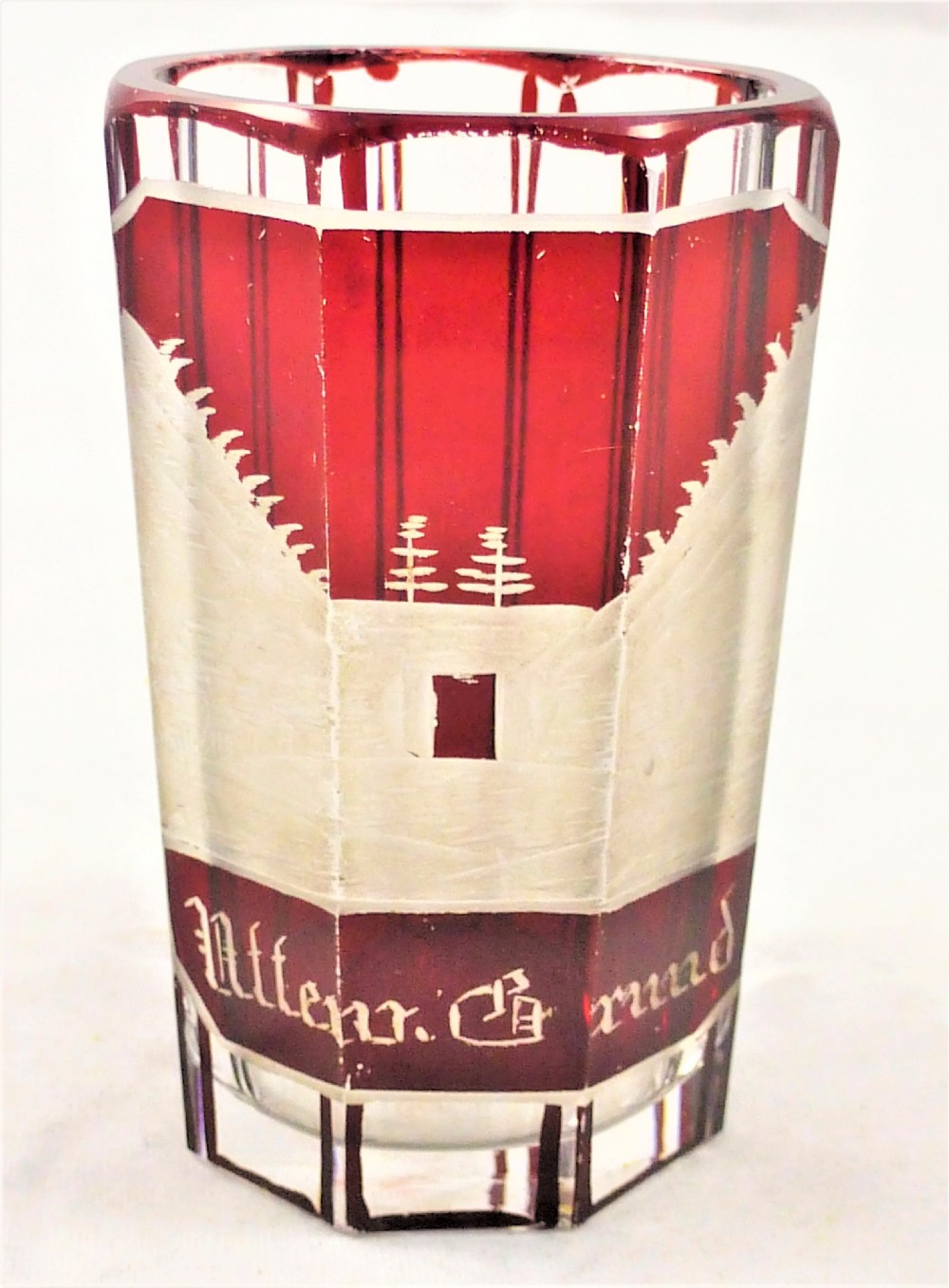 A small faceted glass shot glass featuring a ruby red flashed coating etched with a building scene and German script text.