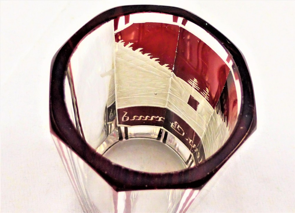 A small faceted glass shot glass featuring a ruby red flashed coating etched with a building scene and German script text. View V