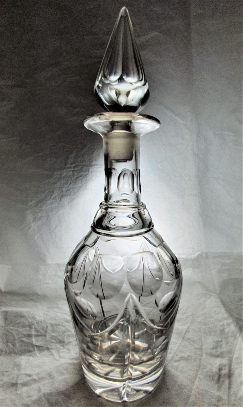 A clear cut lead glass decanter featuring a pointed faceted stopper, cut neck detail, and a carved pattern on the rounded body.