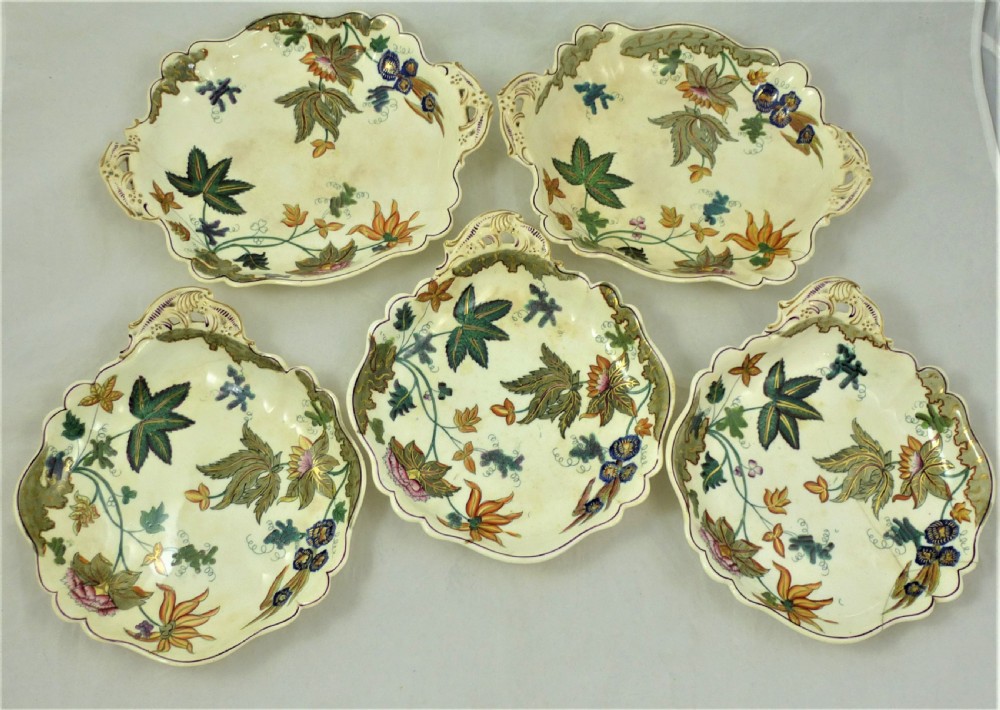 A five-piece English Rococo porcelain dessert service featuring cream-coloured shaped dishes hand-painted with green foliage, floral motifs, and gilt scalloped rims.
