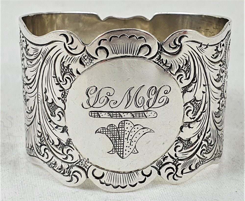 A Victorian silver napkin ring featuring a scalloped rim, detailed scroll engraving, and a central circular cartouche with a monogram.