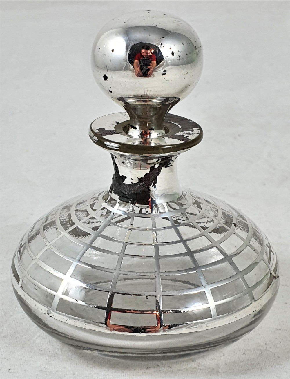 A round clear glass perfume bottle featuring a grid pattern silver overlay and a spherical silver stopper.