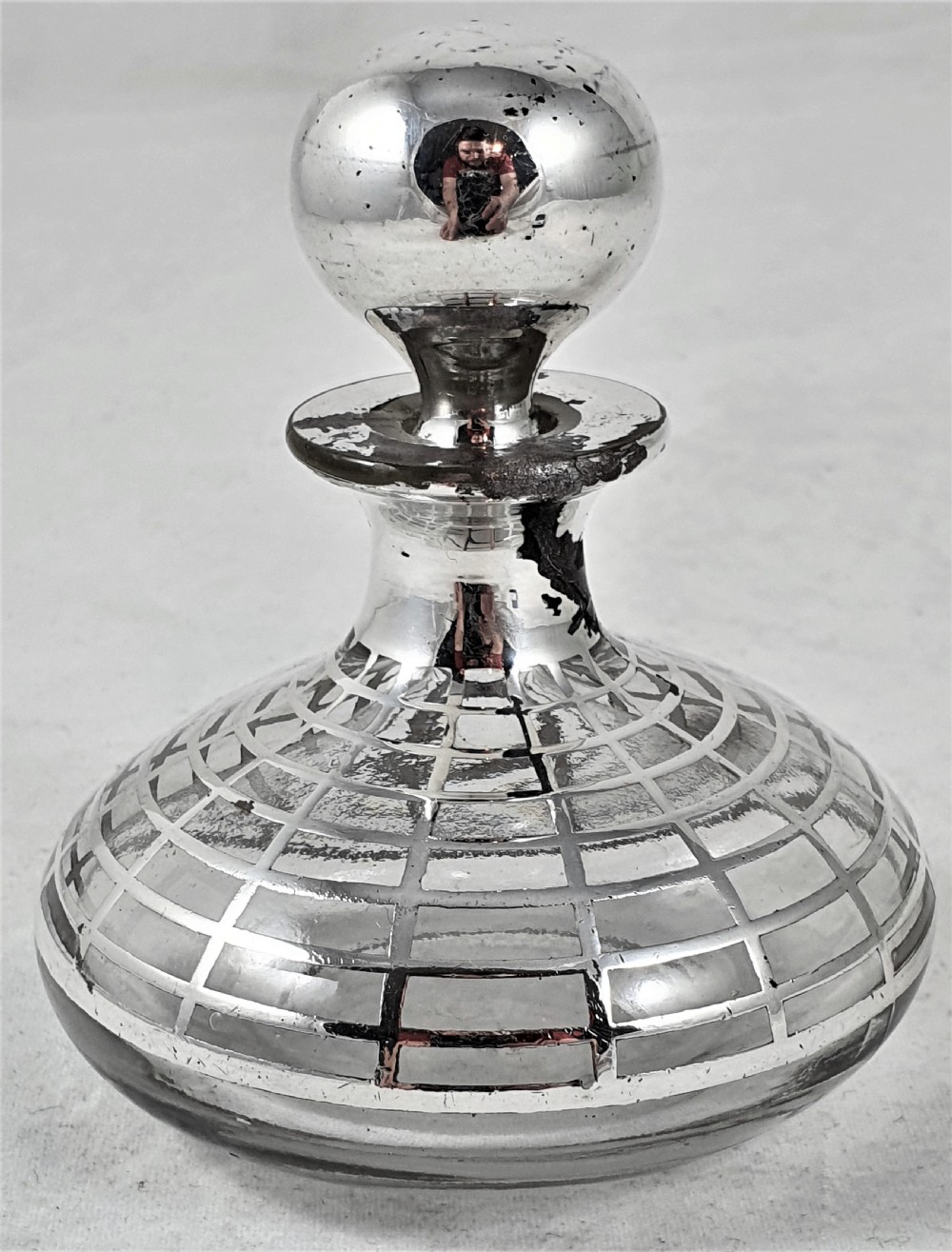 A round clear glass perfume bottle featuring a grid pattern silver overlay and a spherical silver stopper. View II