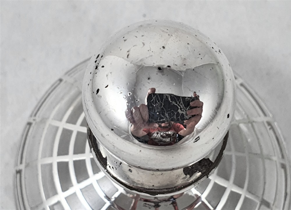 A round clear glass perfume bottle featuring a grid pattern silver overlay and a spherical silver stopper. View V