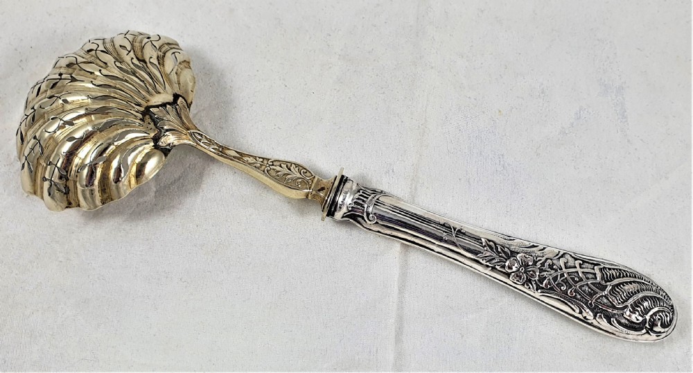 A nineteenth-century French silver-plated sugar sifter featuring a pierced shell-shaped gilded bowl and relief handle, presented in a dark fitted case. View V